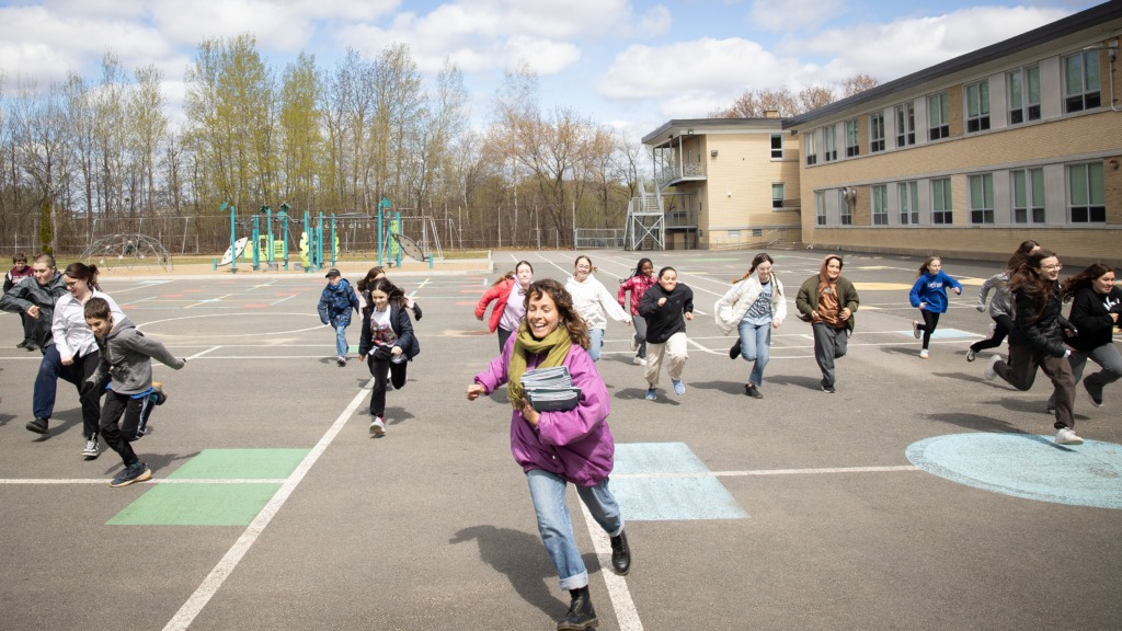 Une adulte court avec des carnets sous le bras, suivie d'un groupe d'élèves dans la cour d'école